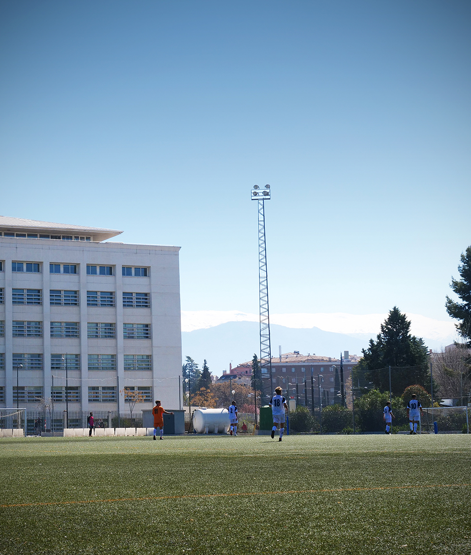 Imagen de un entrenamiento en el campo de fútbol del Campus de Fuentenueva con el edificio del politécnico de fondo, y con un juez de línea y jugadores vestidos de blanco y naranja